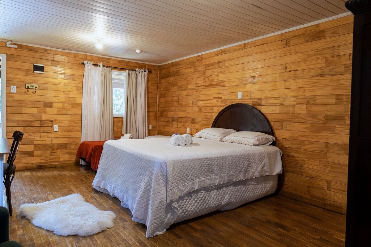 Una habitación con paneles de madera, una cama, una silla, un manto de piel y un ventanal con cortinas.