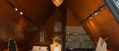 A cozy attic room with wooden walls, a chandelier, a dining table with chairs, a sofa, and a large furry rug.