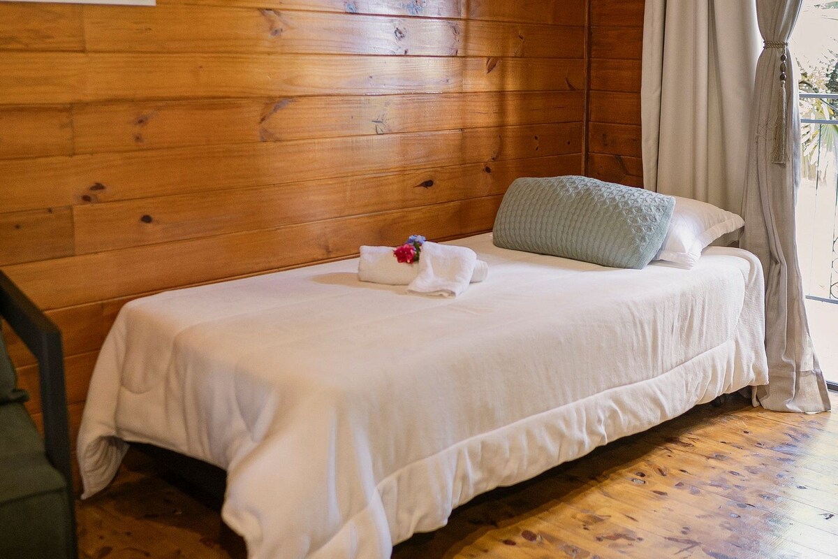 Una habitación tipo cabaña de madera con una cama individual, una silla y una ventana con cortinas.