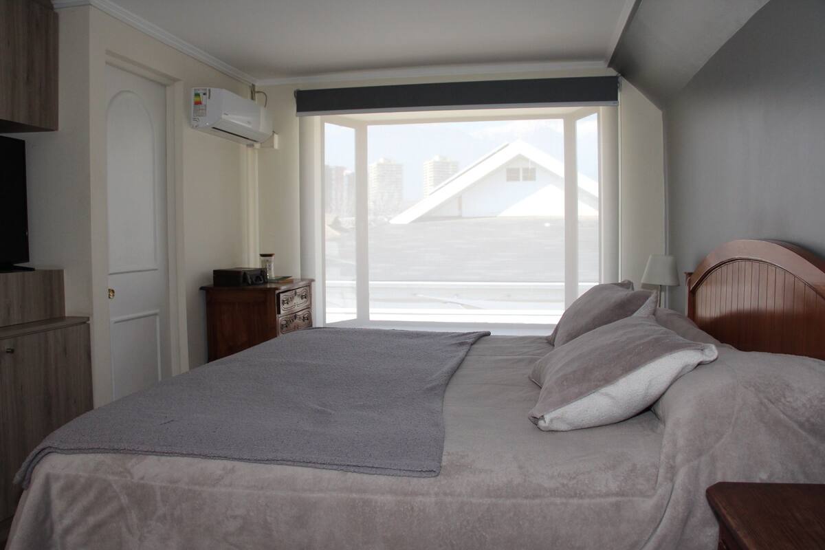 A bedroom with a bed, a television, a dresser, and a window with a view of buildings.