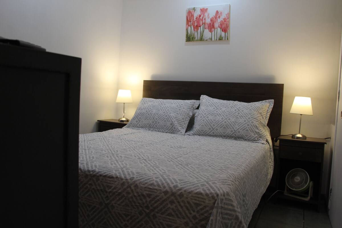 A neatly made bed with patterned bedding, two bedside lamps, a wall-mounted artwork, and a small fan on the nightstand.
