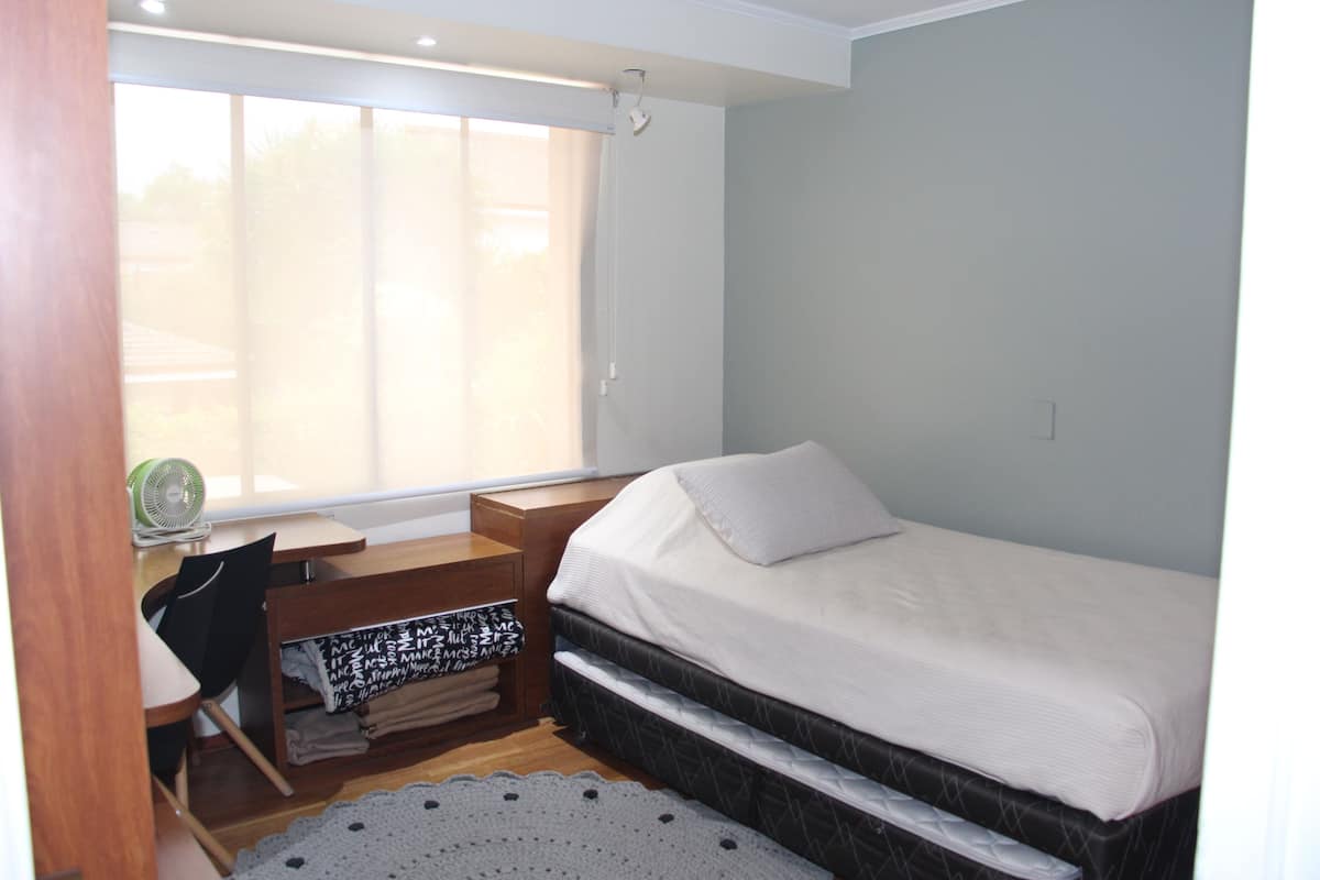 A bedroom with a bed, a desk with a chair, a window, and a small fan.