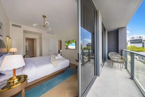 Ein Hotelzimmer mit Bett, Nachttisch, Lampe, Blick auf den Strand durch ein Fenster und einem Balkon mit Stühlen.