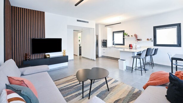 Una sala moderna con televisor de pantalla plana, sofá, mesa de centro y una zona de cocina con mesa de comedor y sillas.