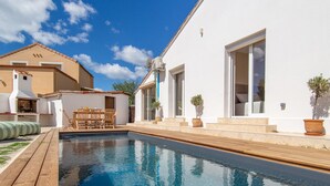 A swimming pool with a wooden deck, a barbecue area, and a white building with large windows.