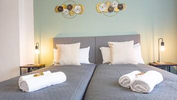 A double bed with a quilted grey bedspread, two white pillows, and two rolled towels on each side.