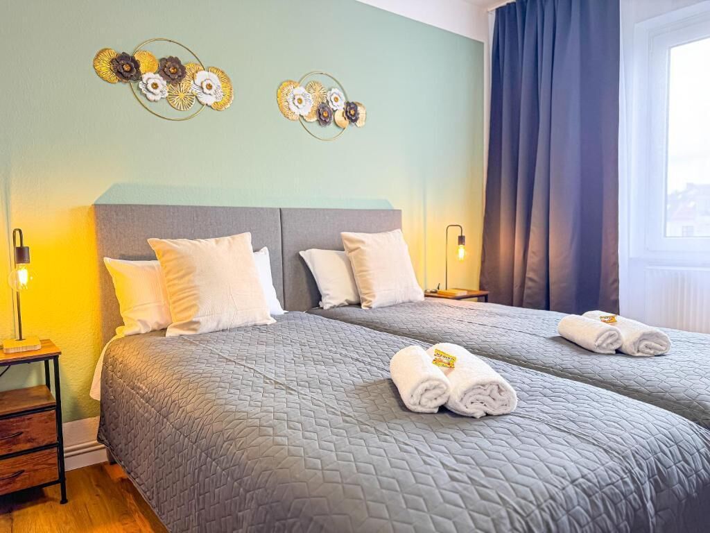 Een hotelkamer met twee bedden, een nachtkastje met een lamp, decoratieve wandkunst en uitzicht op de stad door het raam.