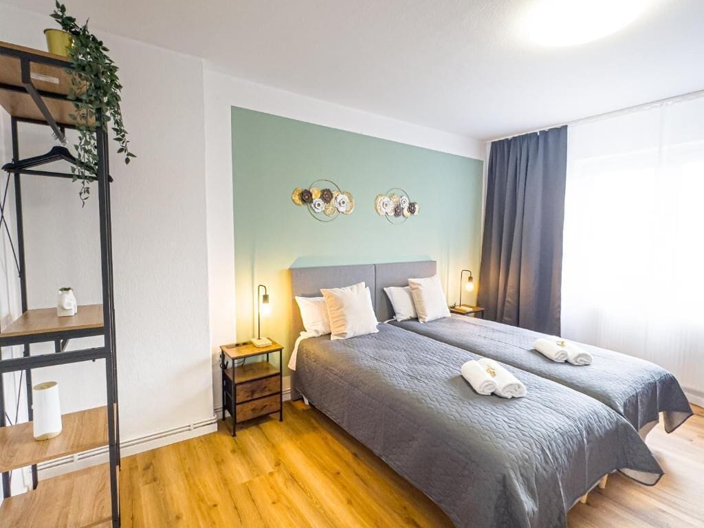A hotel room with two beds, a wooden floor, a green accent wall, and a plant on a shelf.