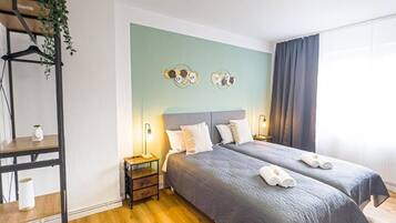 A hotel room with two beds, a wooden floor, a green accent wall, and a plant on a shelf.