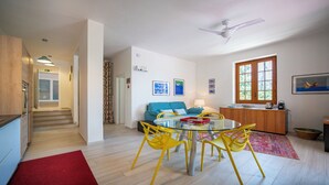 Een moderne woonkamer met een bank, een glazen tafel met gele stoelen, een rood kleed en een keukenhoek met een raam.
