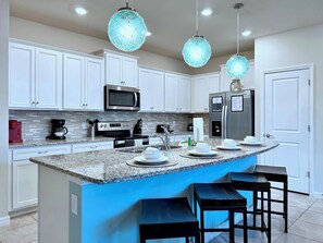 Una cocina moderna con armarios blancos, electrodomésticos de acero inoxidable, encimera de granito e iluminación colgante azul.