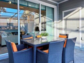 Un área de comedor exterior moderna con una mesa de vidrio y sillas de mimbre, una puerta corrediza de vidrio que da a una sala de estar y vista al cielo.