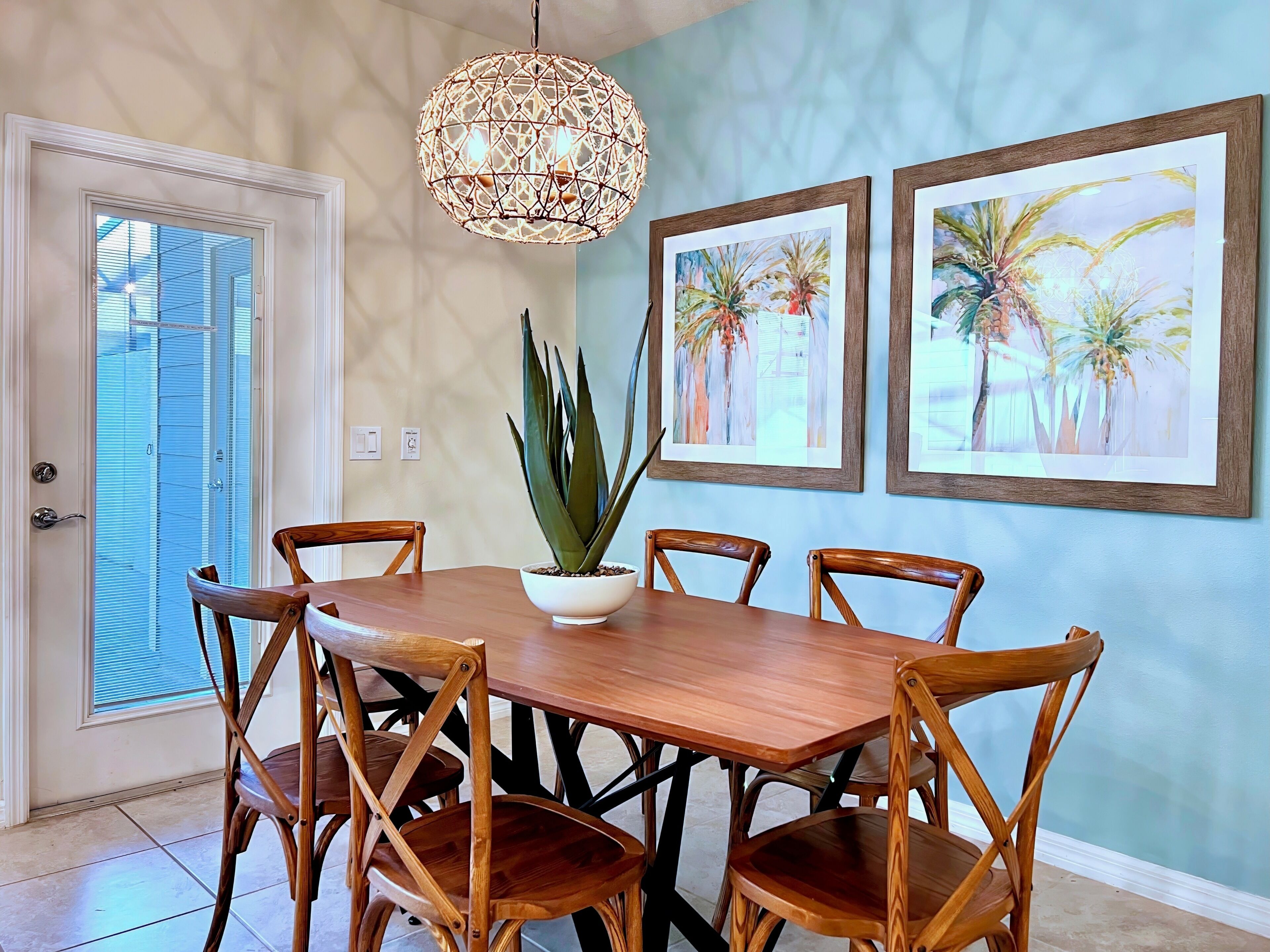 A dining area with a wooden table, six chairs, a potted plant, and two framed paintings on the wall.