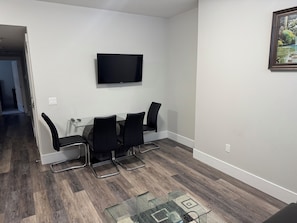 A modern living room with a glass dining table, four black chairs, a flat-screen TV on the wall, and a wooden floor.