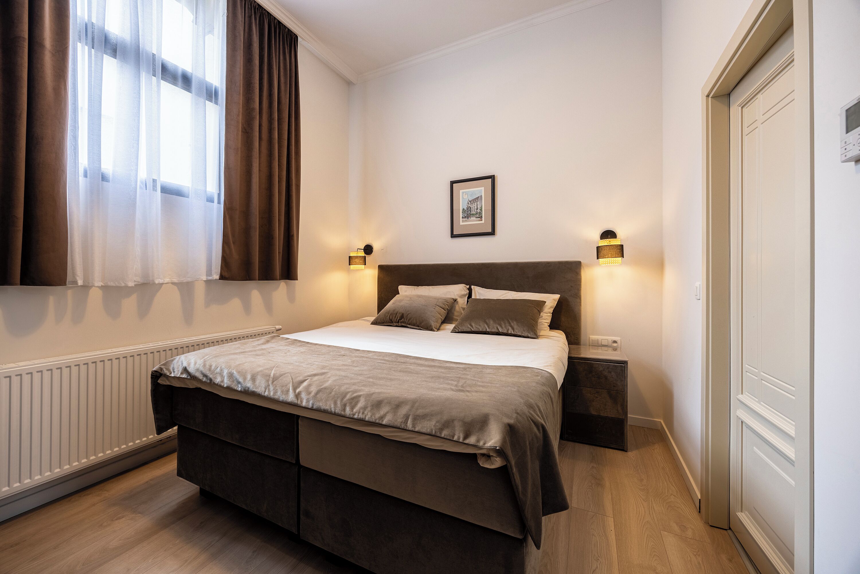 A bedroom with a large bed, two bedside tables, a framed picture on the wall, and a window with curtains.