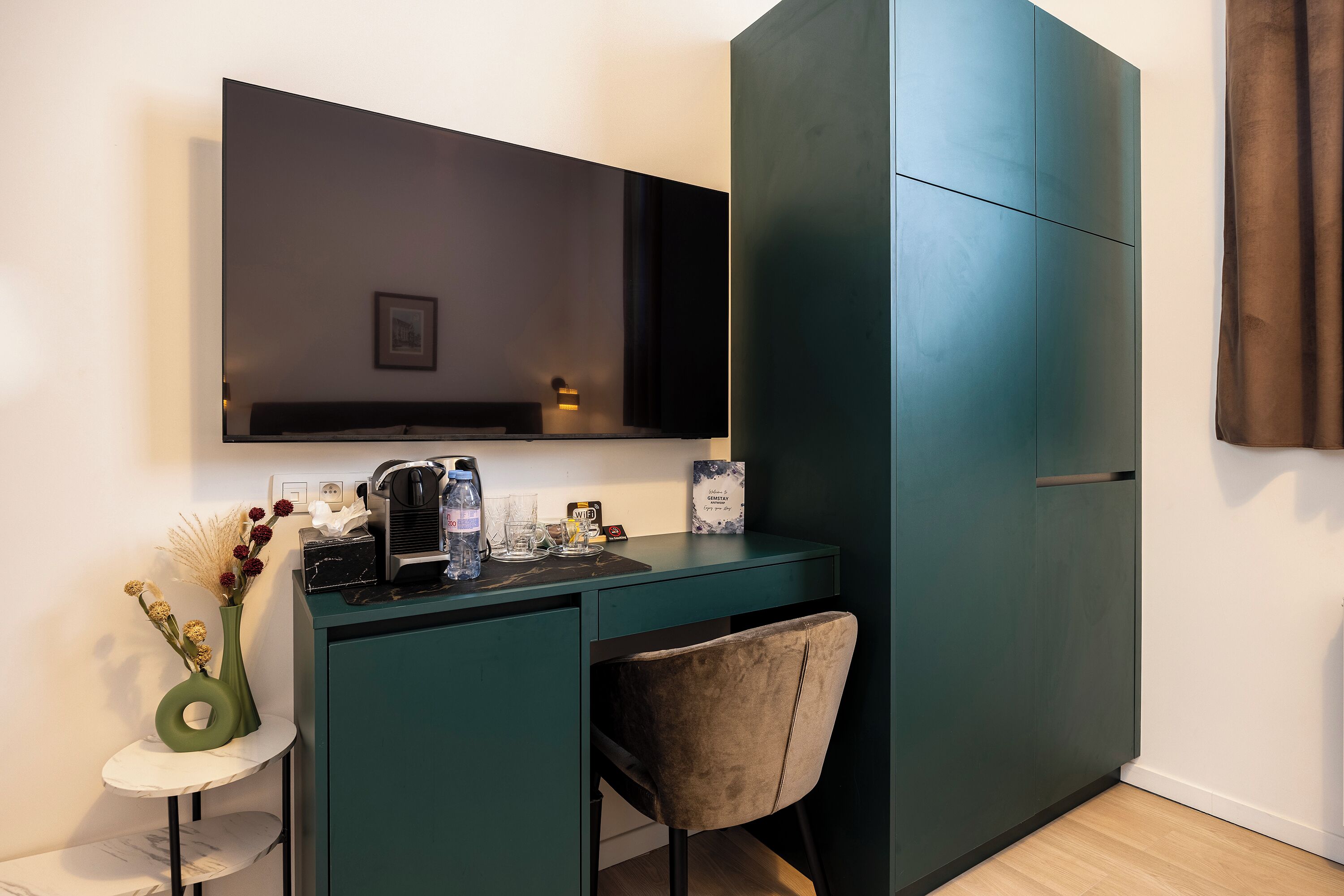 Une chambre moderne avec un bureau vert foncé, un téléviseur fixé au mur, une petite table d’appoint blanche avec un vase, une chaise et une grande armoire.
