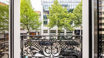 A balcony with ornate metal railings, a view of a modern building with glass windows, and trees in the background.