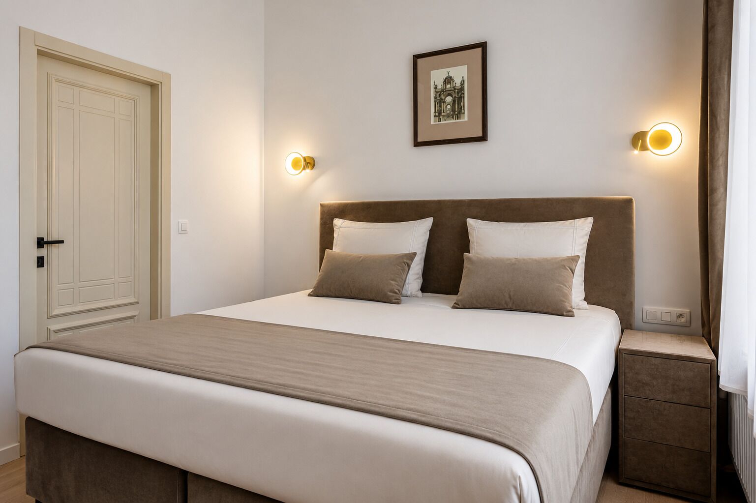 Een net opgemaakt bed met een hoofdeinde, twee nachtkastjes, een deur en een ingelijst beeld aan de muur.