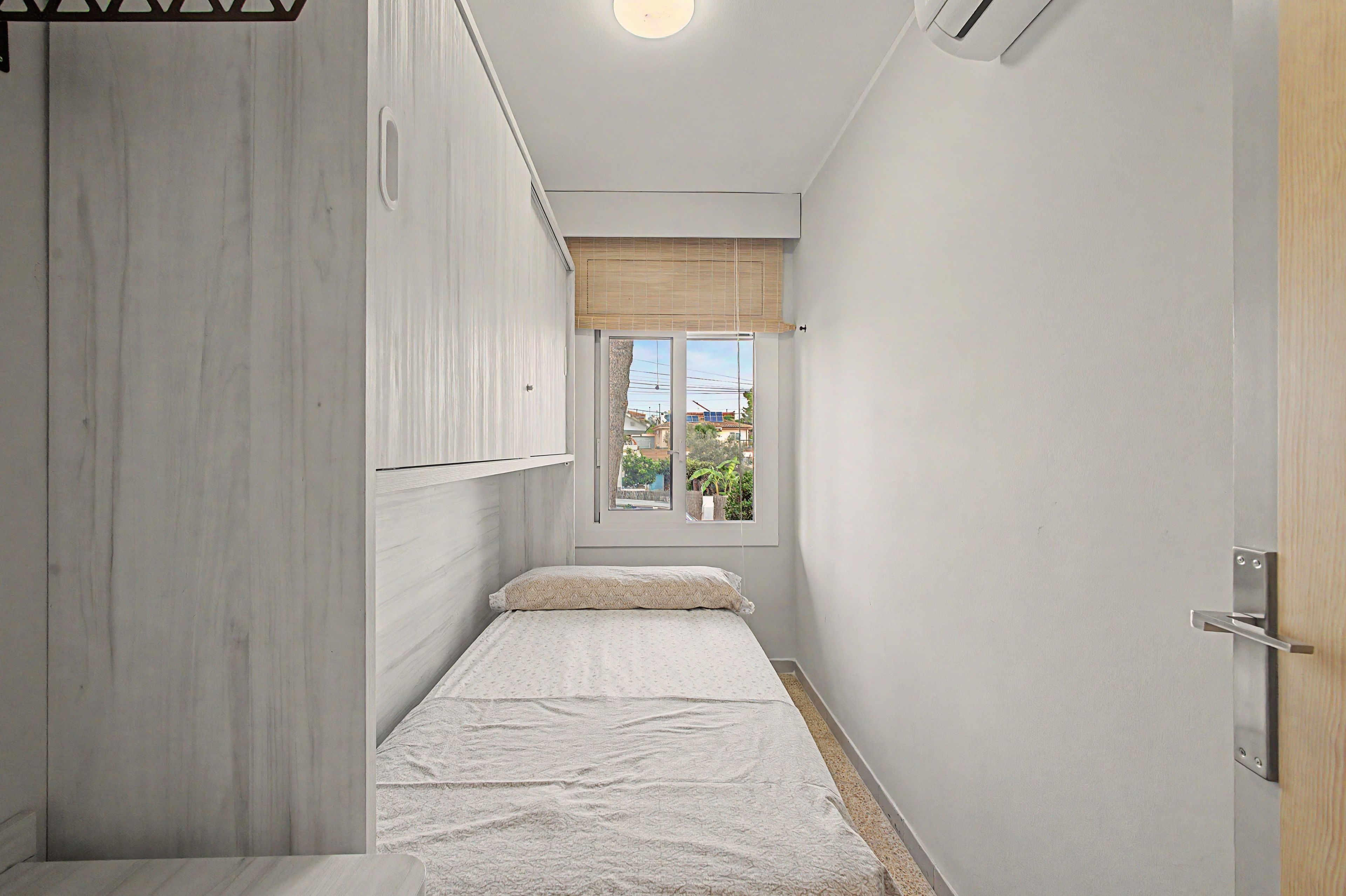 A narrow room with a bed, a window with a view of buildings, and a door.
