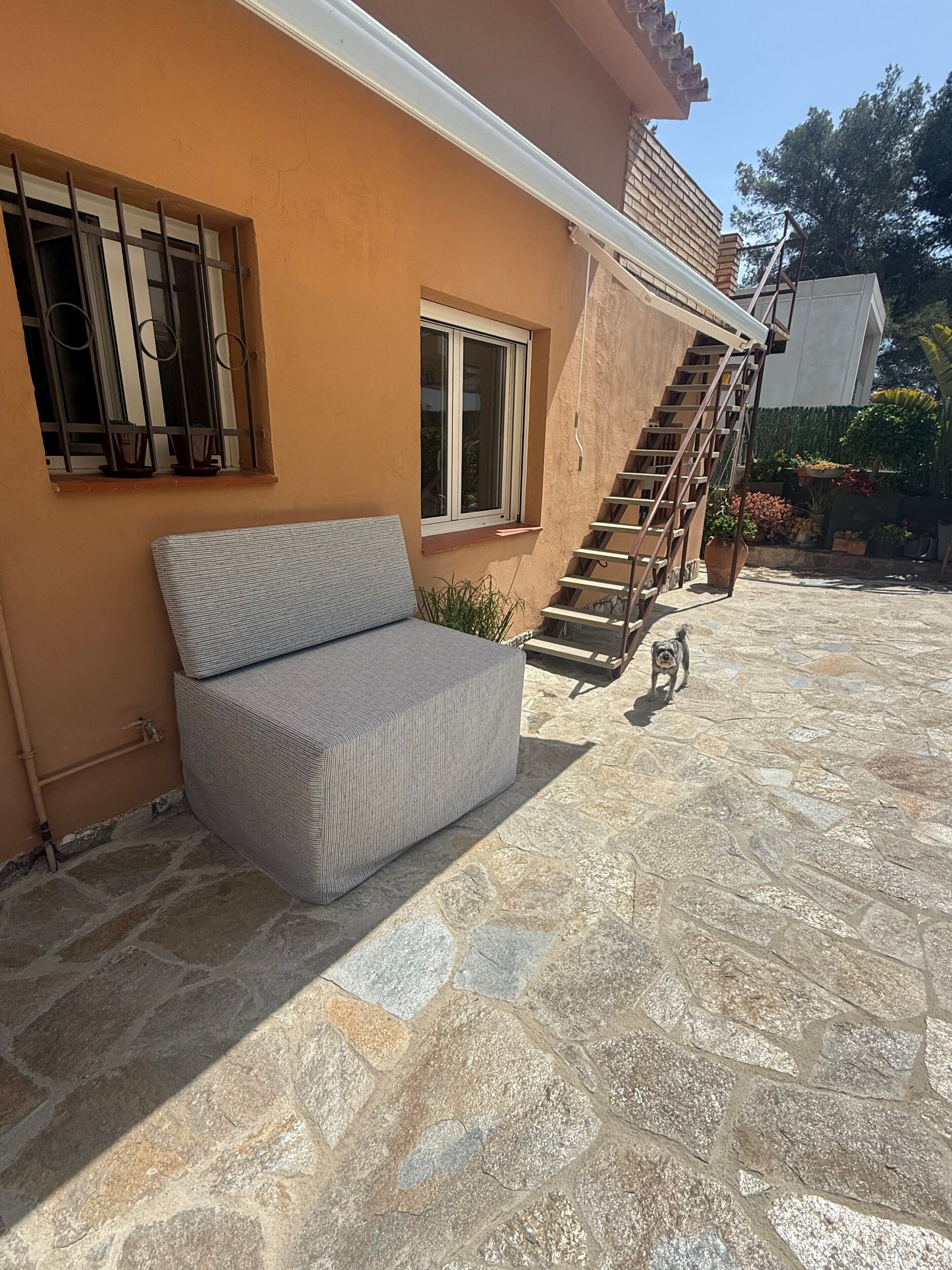 Un patio de piedra con un sofá, una ventana con rejas, una escalera y un perro pequeño.