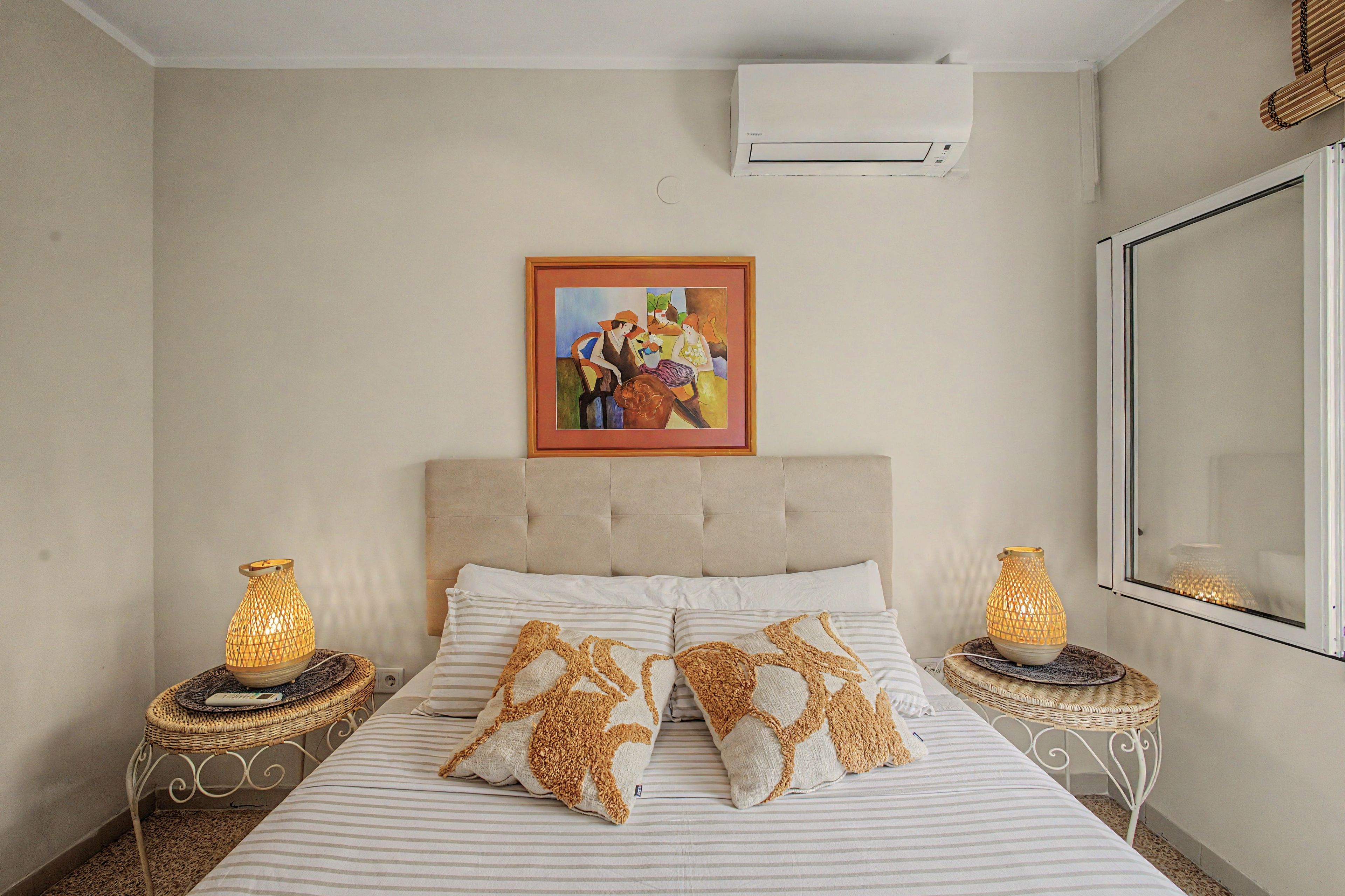 A bedroom with a bed, two nightstands, a wall-mounted air conditioner, and a framed painting.