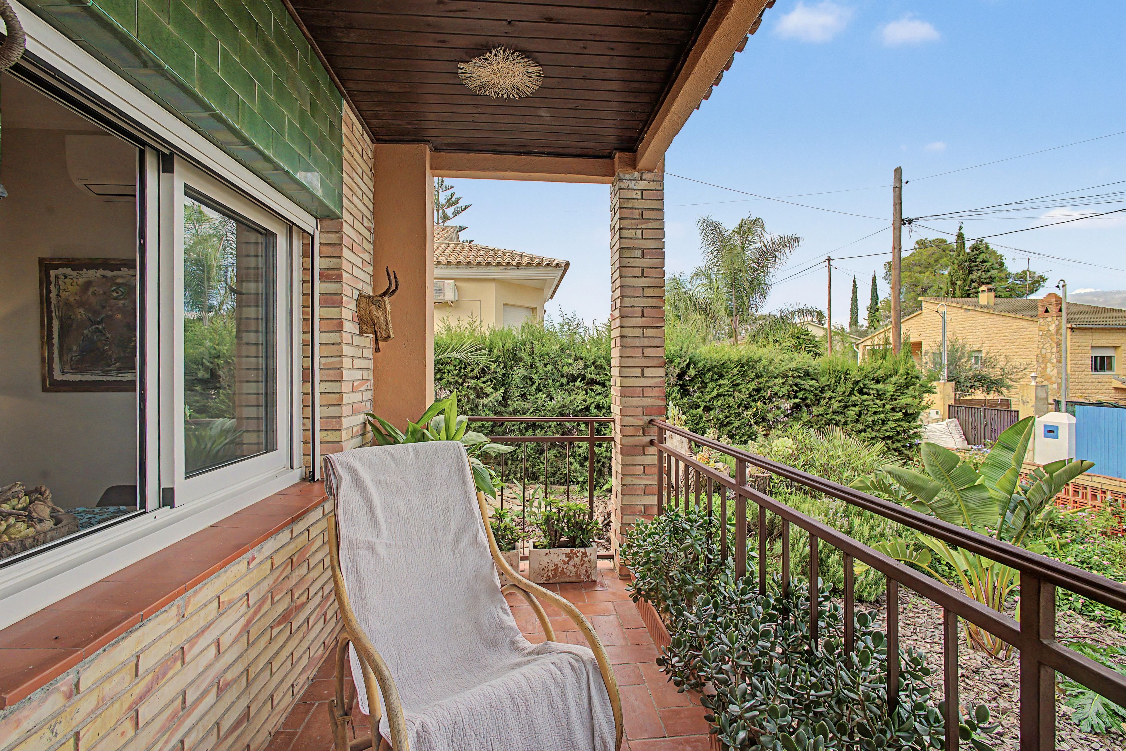 Un balcón techado con una silla, un ventanal y vistas a un jardín y casas vecinas.