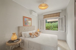 A bedroom with a bed, a painting, a window with a view of greenery, and a hanging light fixture.