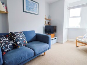 Ein Wohnzimmer mit blauem Sofa, einem Fernseher auf einem Schrank, einem Fenster und einem Bücherregal.
