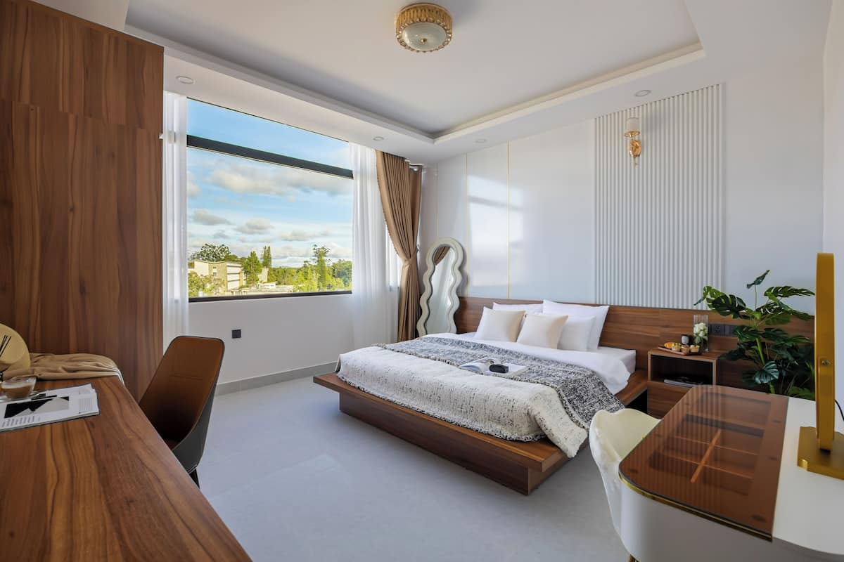 Quarto de hotel moderno com uma cama grande, uma escrivaninha, uma cadeira, mesa de cabeceira, uma planta e vista para o exterior.