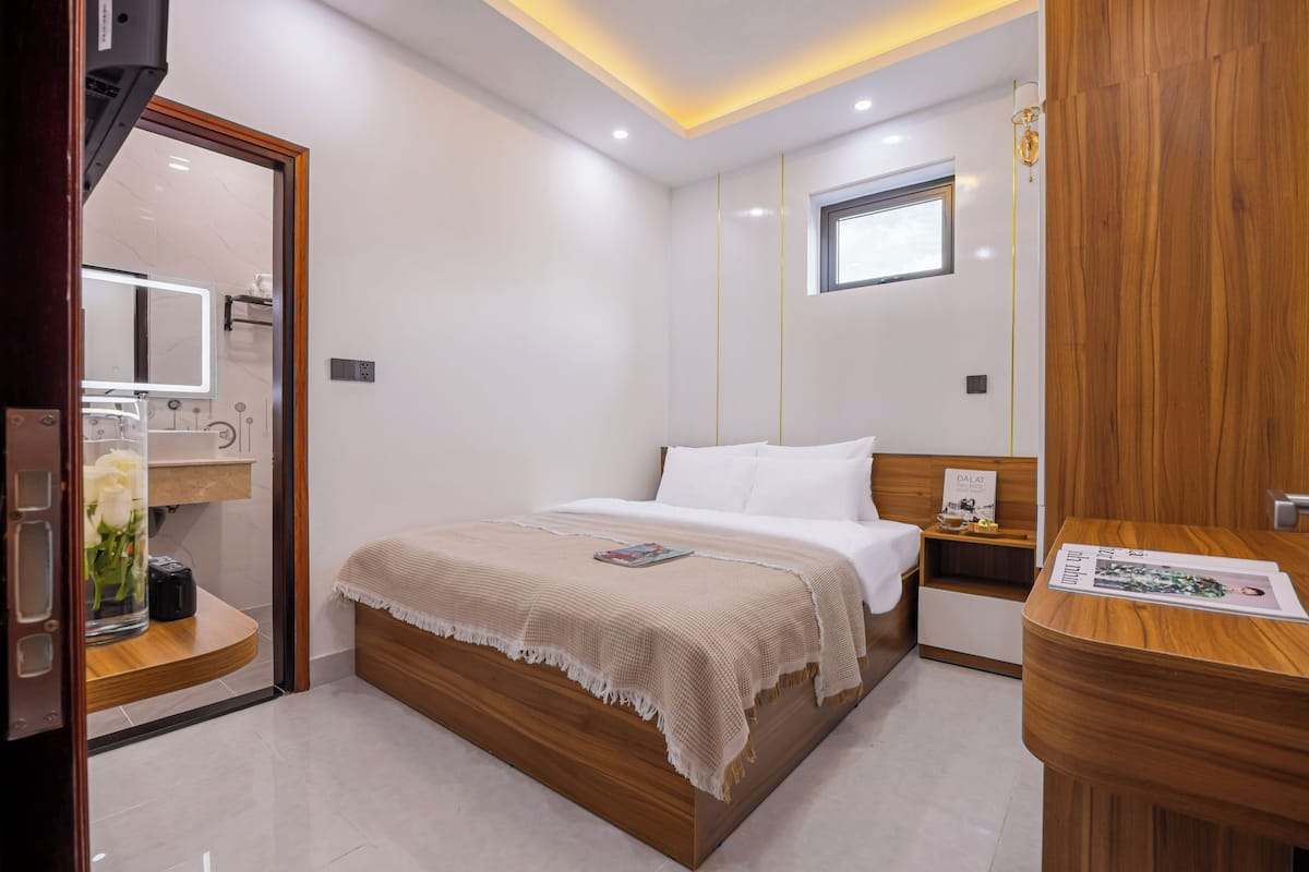 Um quarto moderno com uma cama de madeira, uma mesa de cabeceira, um criado-mudo, um espelho, uma janela e uma pequena prateleira com um vaso de flores.