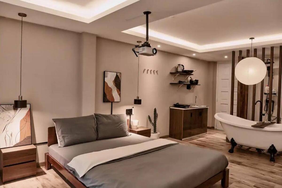 Quarto moderno com cama, mesa de cabeceira, abajur, arte na parede, projetor no teto, armário de madeira, pia, espelho, banheira branca e luminária pendente.