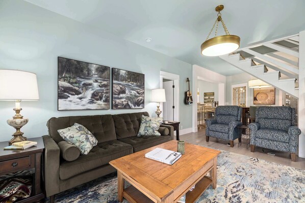 A living room with a sofa, coffee table, and two armchairs. There are paintings on the wall, a staircase, and a guitar. - PLATTinum Haven, In town 3 bedroom 3 bathrooms with hot tub (Red Lodge)