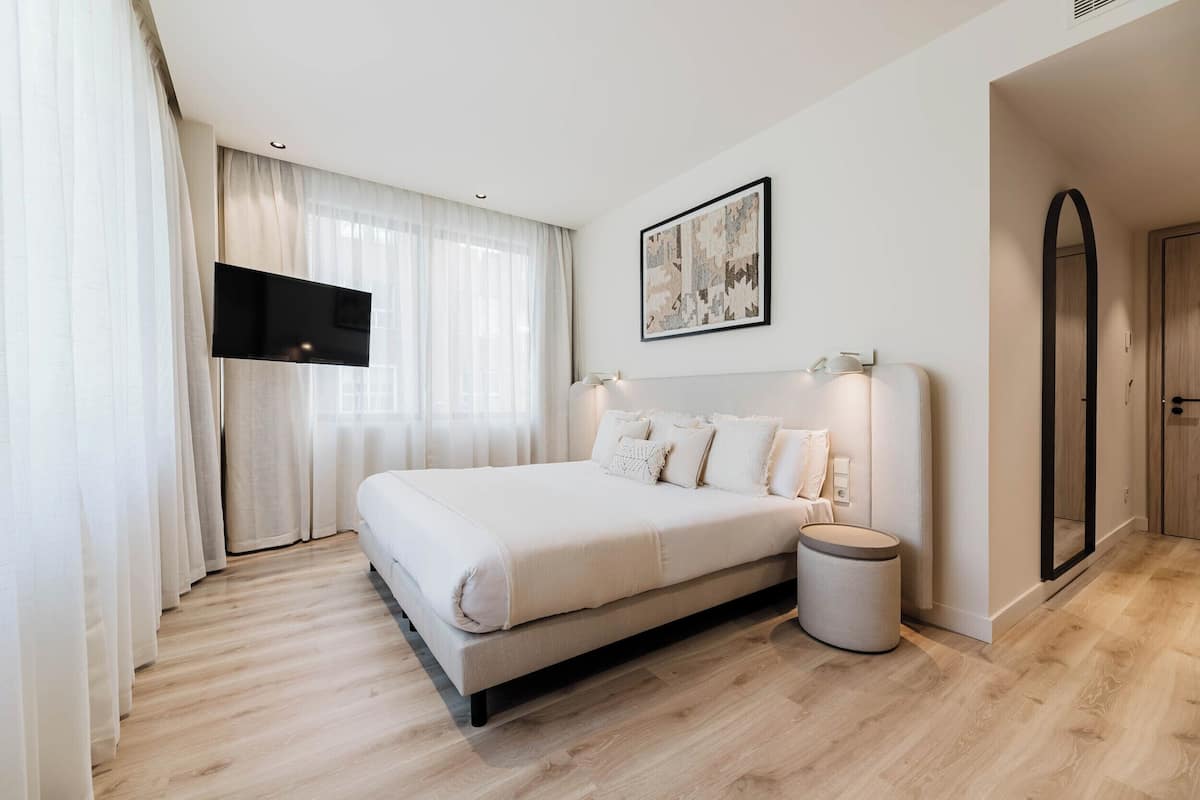 A bedroom with a bed, a TV mounted on the wall, a bedside table with a lamp, and a round trash bin.