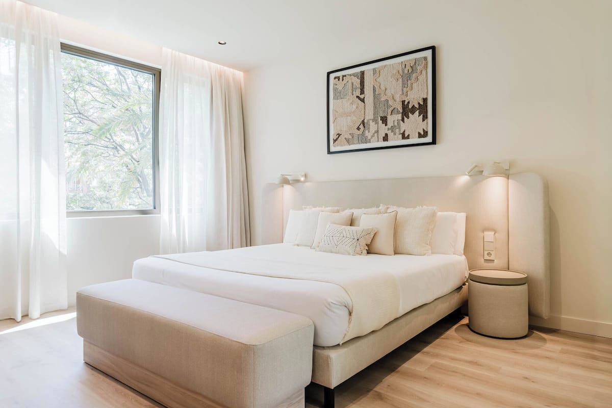 A bedroom with a bed, bedside lamps, a bedside table, a window with curtains, and a wall-mounted artwork.