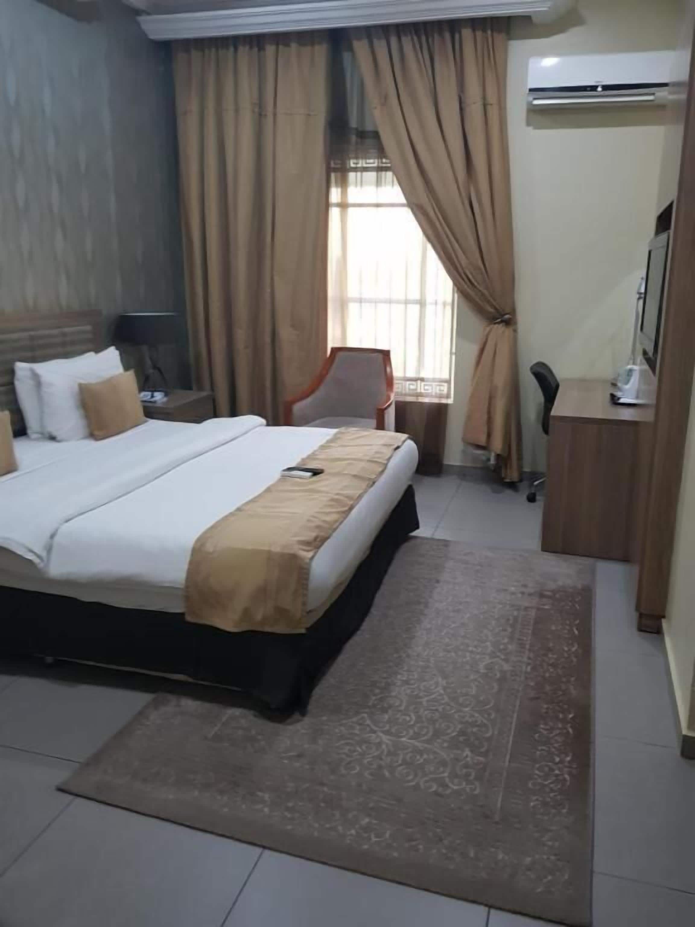 A hotel room with a bed, a desk with a computer, a chair, and an air conditioning unit.