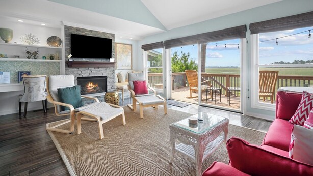 A spacious living room with a fireplace, comfortable seating, and a large sliding glass door leading to an outdoor deck with a view of the countryside. - Spectacular Sunset Views, Quiet Location, Golf Cart Included, Private Deck - 7 Periwinkle Court (Fripp Island)