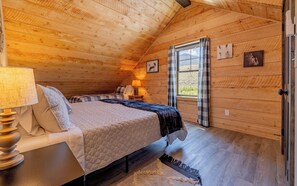 Ein gemütliches Schlafzimmer einer Holzhütte mit Bett, Nachttisch, Lampe, einem Fenster mit Vorhängen und Blick ins Freie.