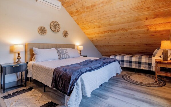 A bedroom with a bed, bedside tables, a lamp, a checkered bedspread, and a wooden ceiling.