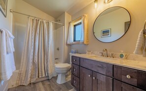 Ein Badezimmer mit Duschvorhang, Toilette, Waschtisch mit Spiegel und einem Fenster.
