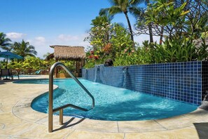 Ein Swimmingpool mit Fliesenrand, ein Gebäude mit Reetdach und üppiger tropischer Vegetation.
