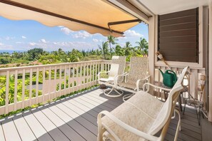 Ein Balkon mit weißen Korbmöbeln, einem grünen Gießkannchen und Blick auf eine tropische Landschaft.