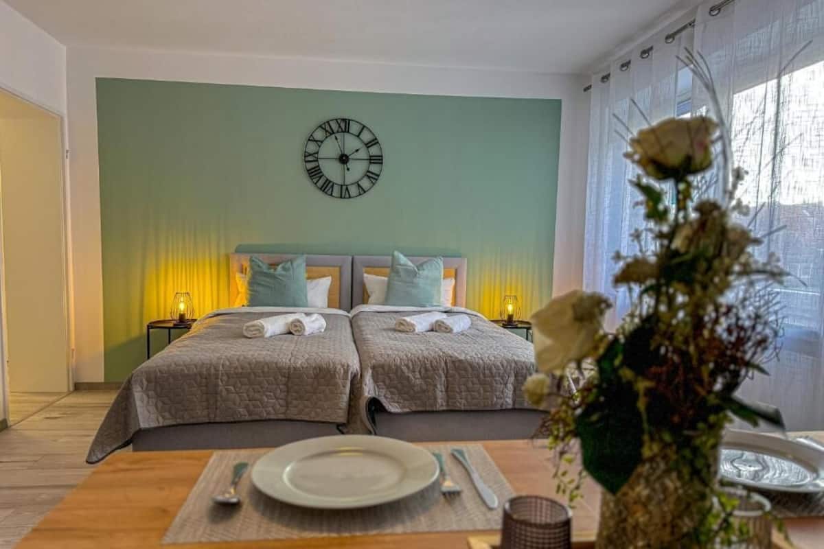 A double room with two beds, a clock on the wall, a dining table with a plate and cutlery, and a vase with flowers.
