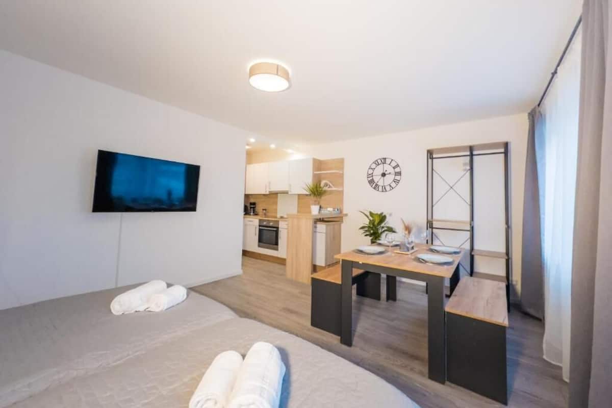A modern hotel room with a bed, a desk, a television, a clock, and a kitchenette.