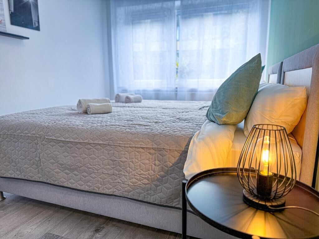 A neatly made bed with a quilted cover, a bedside lamp, and a window with sheer curtains.