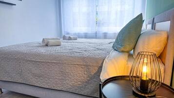 A neatly made bed with a quilted cover, a bedside lamp, and a window with sheer curtains.