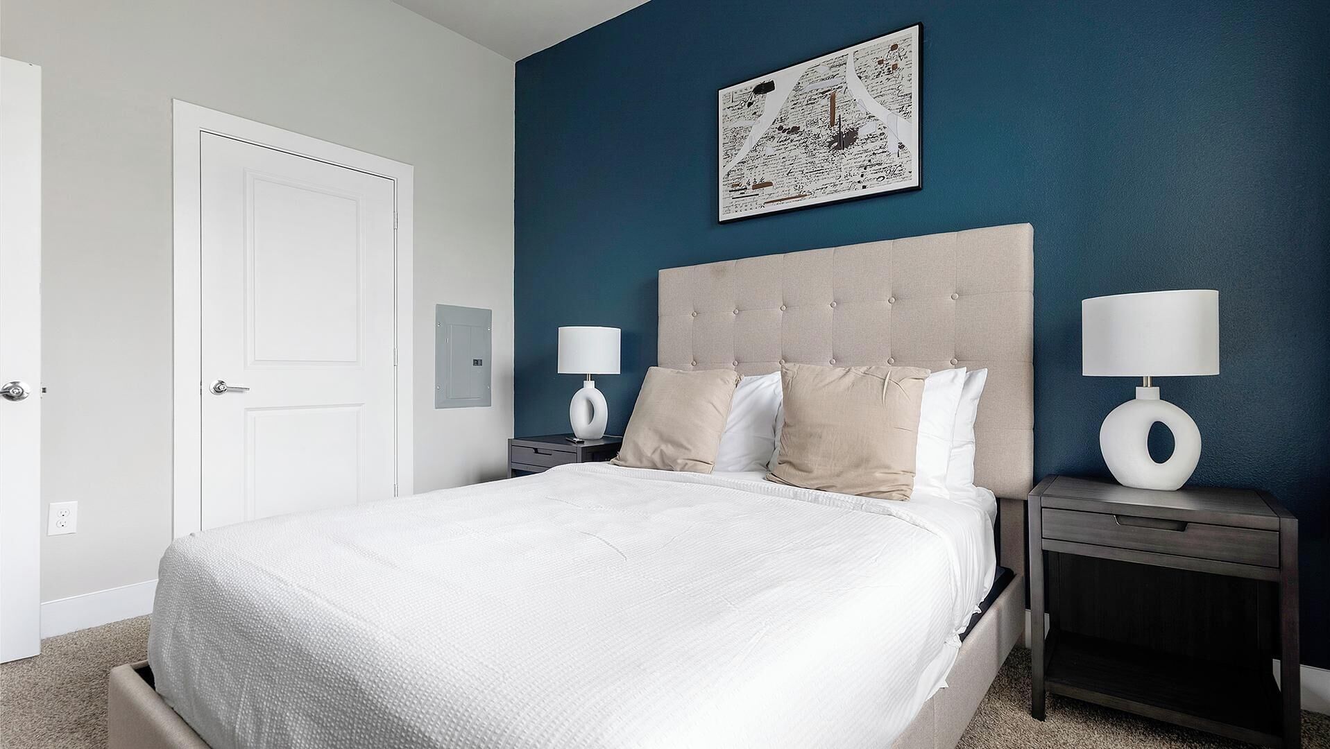 A bedroom with a bed, two bedside tables, a door, and a wall-mounted artwork.
