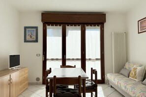 Une pièce comprenant une table à manger, des chaises, un canapé, un téléviseur posé sur un meuble en bois et une fenêtre avec des stores.