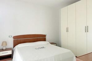 A bedroom with a wooden headboard, a white bedspread, a bedside table with a lamp, and a wardrobe.