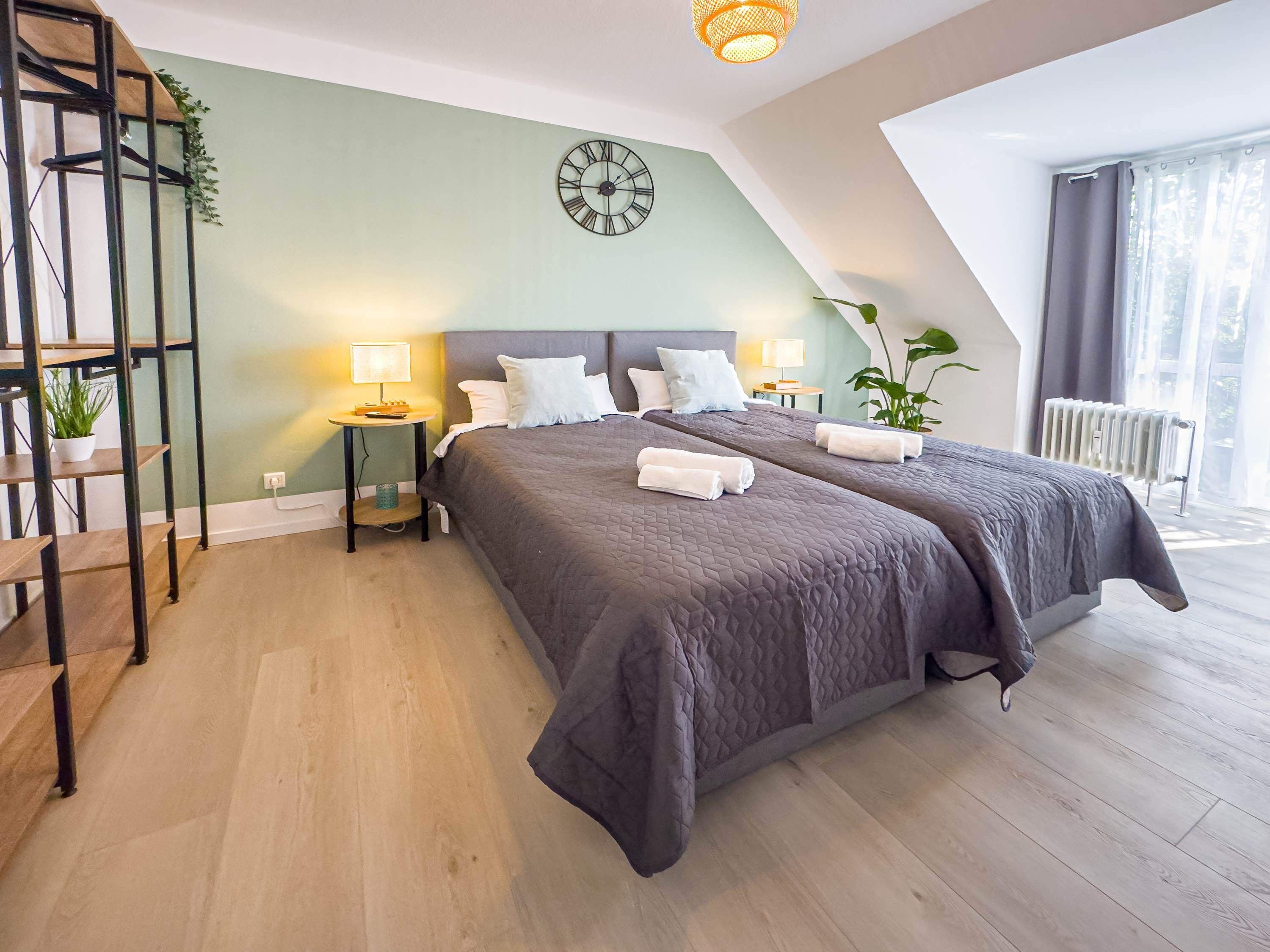 A modern bedroom with a large bed, two bedside tables, a wall clock, and a view of the outdoors.