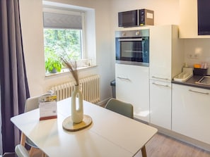 Eine kompakte Küche mit Mikrowelle, Backofen und Kühlschrank, einem Esstisch mit einer Vase sowie ein Fenster mit Blick auf Grünflächen.